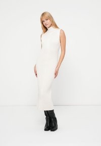 Φόρεμα μέχρι το γόνατο με ριμπ ύφανση σε εκρού χρώμα, με ψηλό γιακά και χωρίς μανίκια. Φοριέται με μαύρες μπότες με τακούνι.