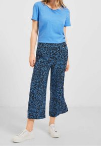 Chemise bleue à manches courtes associée à un pantalon large noir arborant un motif de feuilles bleues. Portée avec des baskets blanches à lacets.