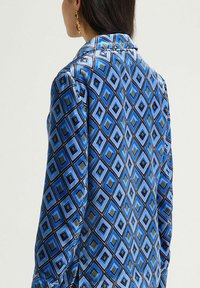 Chemise à manches longues en satin bleu présentant un motif géométrique en forme de losange dans des nuances de bleu, vert et noir ; col tailleur et poignets à boutons.