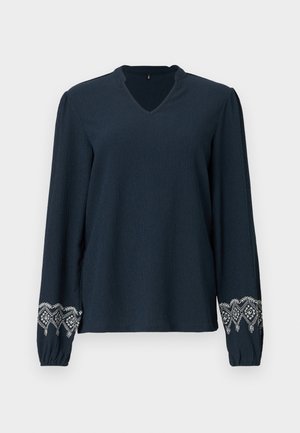 Blusa blu navy con scollo a V, tessuto strutturato e maniche lunghe. Presenta un intricato ricamo bianco sui polsini.