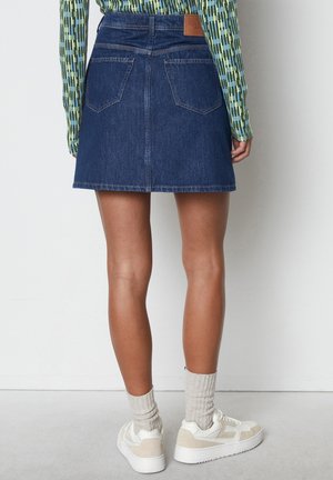 Bagfra af en person iført en blå denimnederdel, mønstret langærmet trøje, lyse beige sokker og hvide sneakers, stående på en lys gulv.