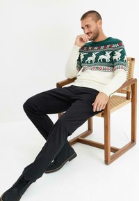 Pullover lavorato a maglia con un motivo verde e crema che presenta renne e disegni floreali, abbinato a pantaloni neri e stivali neri, seduto su una sedia di legno.