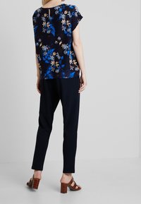 Bluse mit Blumenmuster in Marineblau und kurzen Ärmeln, mit einem Schlüssellochdetail am Rücken. Kombiniert mit marineblauen Hosen und braunen Sandalen mit Blockabsatz.