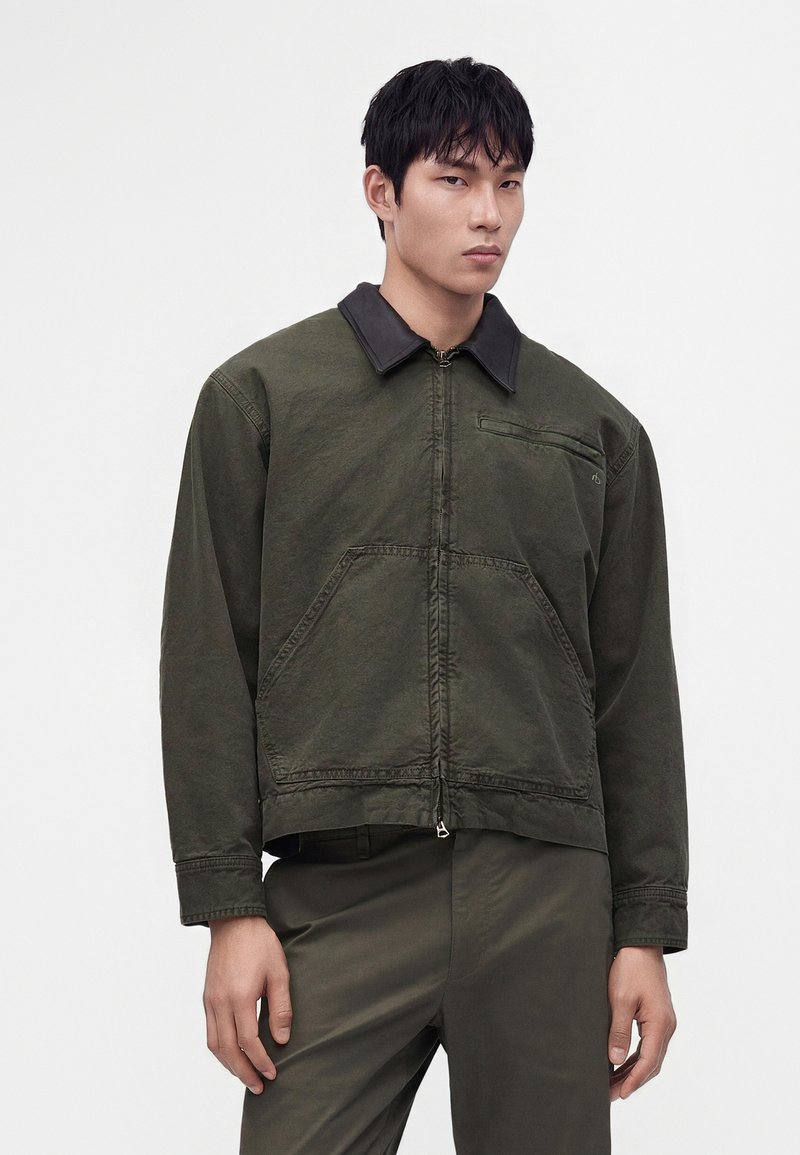 Jeune homme portant une veste zippée vert olive avec de grandes poches avant et un col sombre, assortie à un pantalon vert olive.