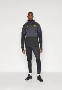 Sort atletikjakke med blå og gule accenter, lynlås i halsen, logo-patch, parret med sorte bukser med hvide side striber og sorte sneakers.