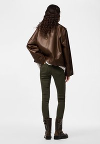 Osoba s dugom kovrčavom kosom koja nosi smeđu kožnu jaknu, maslinasto zelene uske hlače i tamnosmeđe čizme s kopčama, stoji okrenuta leđima.