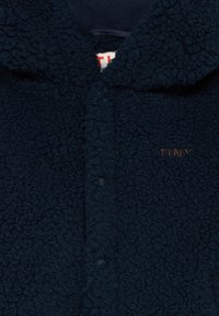 Veste en polaire texturée bleu marine foncé avec col, boutons-pression et petit texte brodé brun "TINY" sur la poitrine.