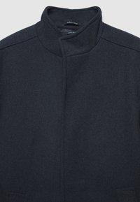 Cappotto in misto lana scuro con colletto alto, forma strutturata e design minimalista. Presenta chiusura frontale nascosta e tasche laterali.