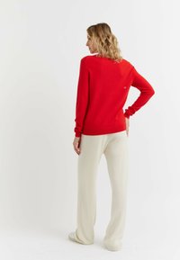 Rode trui met lange mouwen, ronde hals en subtiele logodetail op de achterkant, gecombineerd met crèmekleurige broek met brede pijpen en lichte schoenen.