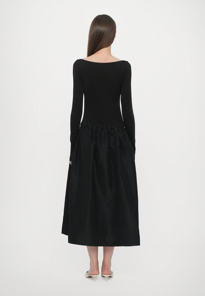 Vestido negro con un cuerpo ajustado de manga larga y una falda de seda volumétrica y recogida. Textura suave con un detalle de lazo en la parte posterior.