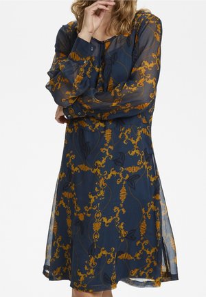 Vrouw draagt een doorschijnende marineblauwe jurk met lange mouwen met een sierlijk goud-zwart bloemmotief, met knopen op de manchetten en aan de voorkant.