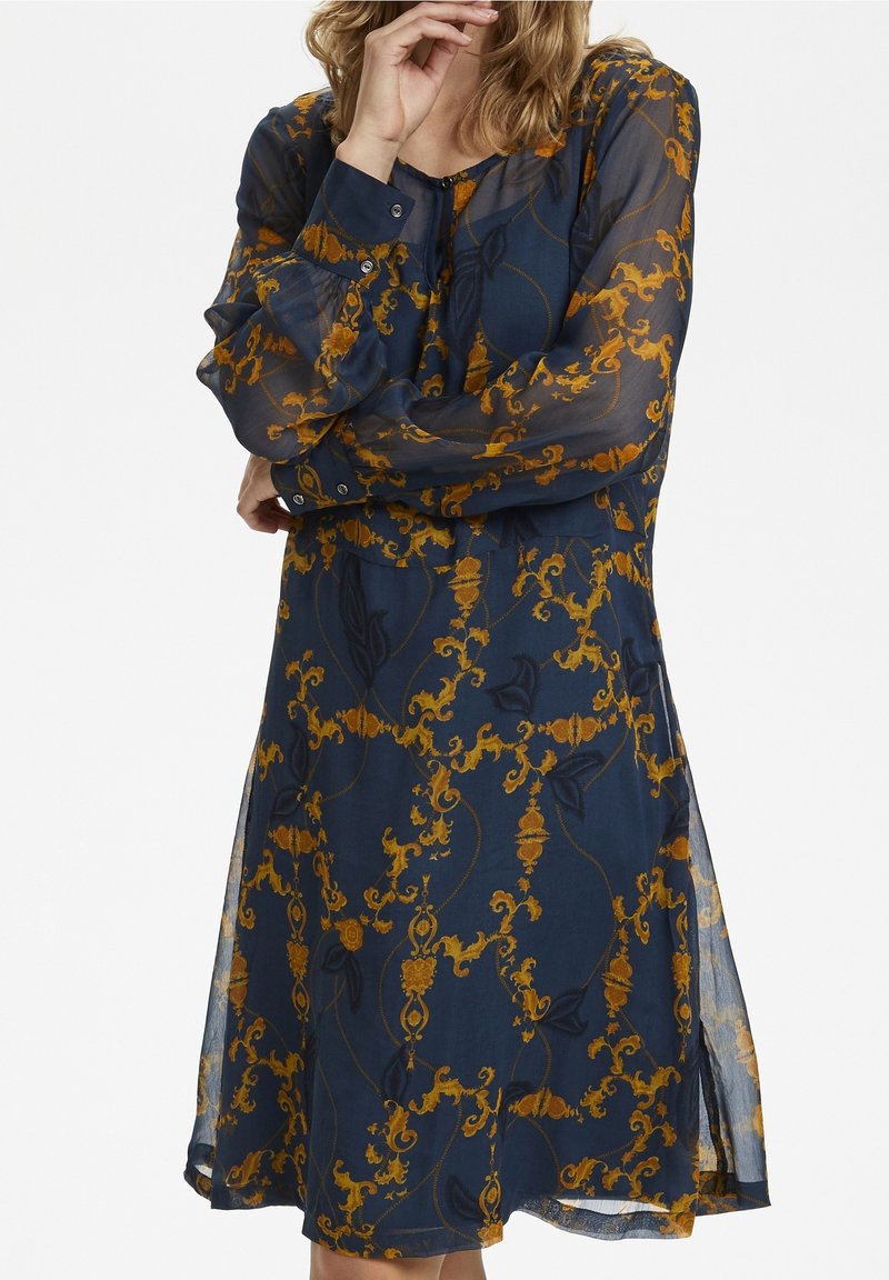 Femme portant une robe bleu marine transparente à manches longues présentant un motif floral orné doré et noir, avec des boutons aux poignets et à l'avant.