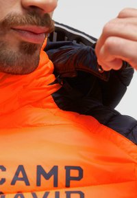 Orange and navy puffer jacket with logo, showcasing a quilted texture and visible zippers. Partially zipper opened at the collar.