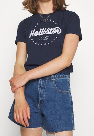 Marineblauwe katoenen T-shirt met witte opdruk waarop "Hollister" staat, en denim shorts met rafelige zomen, voorzien van voorzakken en een knoopsluiting.