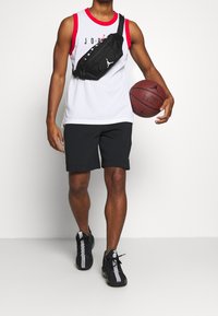 Vit mesh jersey-topp med röda detaljer, svarta shorts, svart midjeväska med logotyp, håller en basketboll, bär svarta sportskor.