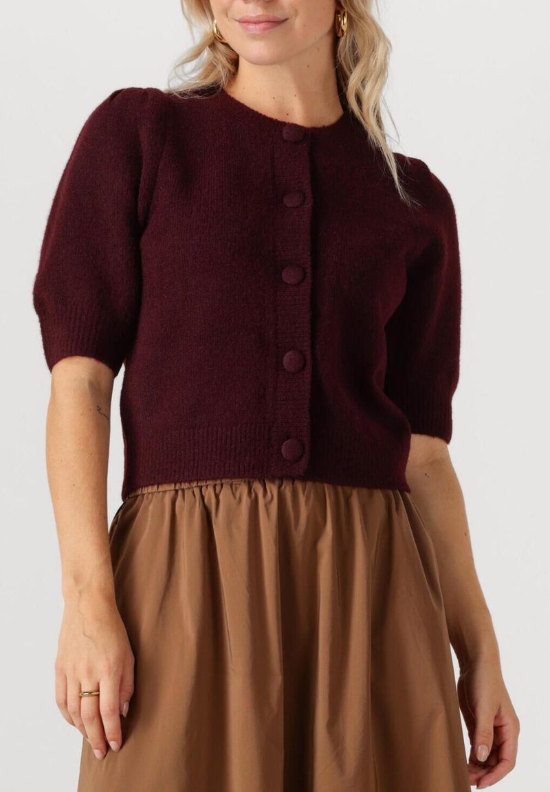 Bordeaux kleurige gebreide cardigan met korte mouwen, ronde halslijn, vijf grote met stof bedekte knoppen en een cropped zoom, gecombineerd met een bruine rok.