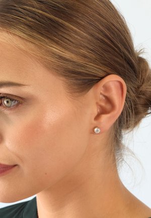 Goldene Steckerohrringe mit einem klaren runden Edelstein, in einem klassischen Design, vor einem glatten, neutralen Hintergrund präsentiert.