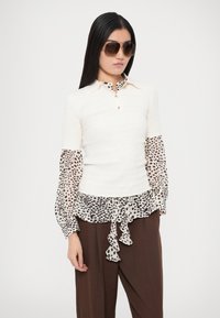 Biela rebrovaná blúzka s dlhými rukávmi s priehľadnými rukávmi a lemom so vzorom leopardieho tigríka, s golierom a detailom s volánmi a mašľou. Hnedé široké nohavice a slnečné okuliare.