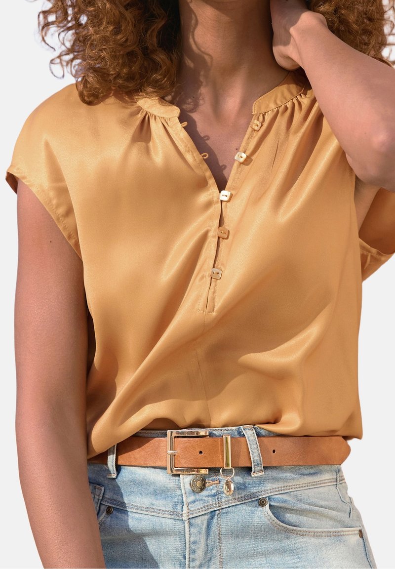 Femme aux cheveux bouclés portant un chemisier jaune moutarde à manches courtes et un jean bleu clair avec une ceinture en cuir marron.