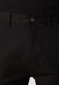 Black jeans with a smooth texture, featuring a front button closure, belt loops, and subtle stitching details. Brand logo visible on button.