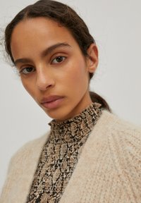Beige textuur cardigan over een zwart gemusterd high neck top, met een close-up van het gezicht met natuurlijke makeup en los haar.