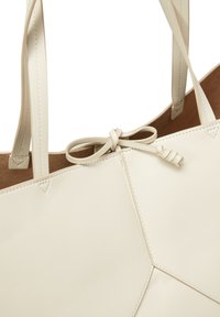 Sac en cuir crème avec une surface texturée, des coutures géométriques et un détail de nœud central. Fines bretelles assorties.