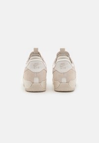 Beige suede sneakers with white accents, featuring a textured sole and heel pull tab. Logo displayed on the back. Rounded shape.