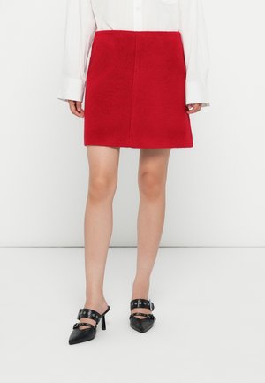 Femme portant une mini-jupe rouge, un chemisier blanc à manches longues et des chaussures à talons hauts noires à bouts pointus avec des brides à boucle.