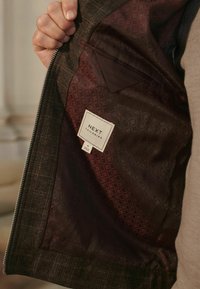 Tailored jacket with a textured, dark brown exterior and a patterned maroon interior. Features a sewn label with "NEXT TAILORING."