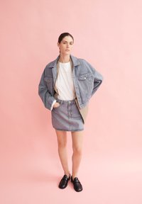 Veste en denim bleu clair, avec une coupe décontractée et deux poches. Assortie à un T-shirt blanc et une jupe en denim assortie. Chaussures noires à enfiler.