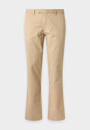 Un homme porte une casquette et un pull bleu marine, une chemise bleu clair à col, un pantalon beige et des bottes marron, sur un fond gris uni.