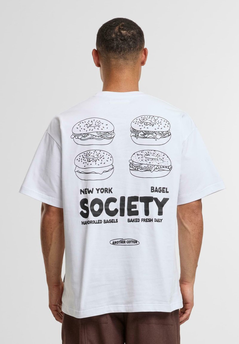 Balta kokvilnas T-kreklā ir grafiska druka ar četriem beigu maizēm un uzraksts "NEW YORK BAGEL SOCIETY" trešajā melnā burtā.