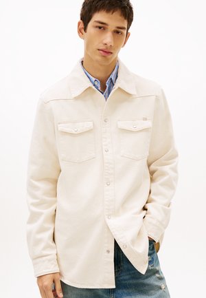 Mladý muž v krémovej zapínacej bunde cez modrú pruhovanú košeľu a potrhané modré rifle, stojaci s jednou rukou v vrecku.