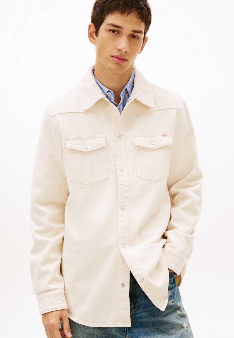 Νέος άνδρας που φοράει ένα κρεμ σακάκι με κουμπιά πάνω από μπλε ριγωτό πουκάμισο και φθαρμένο μπλε τζιν, στέκεται με το ένα χέρι στην τσέπη.