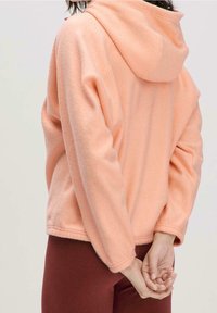 Felpa con cappuccio in pile color pesca dalla texture morbida, taglio oversize e cappuccio con coulisse, abbinata a leggings color bordeaux. Vista posteriore con mani intrecciate.