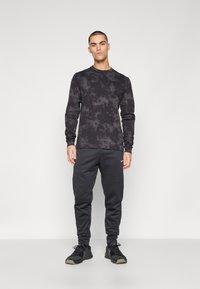 Black long-sleeve shirt with tie-dye pattern, paired with black sweatpants featuring a small white logo. Model wears black athletic shoes.