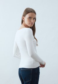 Top bianco a maniche lunghe con scollo rotondo, design aderente e tessuto testurizzato abbinato a jeans blu scuro. Sfondo semplice.