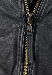 Detail van een zwarte leren jas met een messing rits en een leren trekkoord, waarbij de gestructureerde leather en contrasterende stiksels worden getoond.