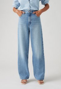 Person wearing light blue high-waist wide-leg jeans with hands in pockets, light blue button-up shirt, and beige open-toe sandals.