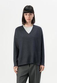 Maglione a v grigio scuro in un morbido tessuto a maglia, dotato di maniche lunghe e una vestibilità rilassata, indossato sopra una t-shirt grigio chiaro.