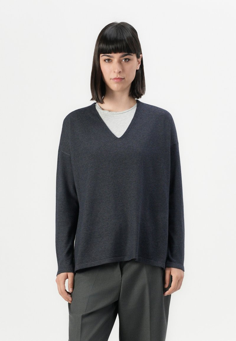 Maglione a v grigio scuro in un morbido tessuto a maglia, dotato di maniche lunghe e una vestibilità rilassata, indossato sopra una t-shirt grigio chiaro.
