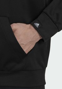 Felpa nera con tasca a marsupio, caratterizzata da un polsino con etichetta cucita del logo Adidas e tessuto strutturato. Mano appoggiata nella tasca.