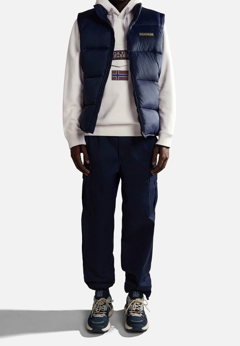 Granatowa kamizelka puchowa na jasnym hoodie z grafiką, zestawiona z granatowymi spodniami cargo i ciemnymi sneakersami. Elementy wyróżniające to zamki błyskawiczne i kieszenie.