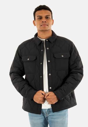 Chaqueta negra acolchada con cierre de botones a presión, dos bolsillos frontales y un cuello estructurado, llevada sobre una camisa blanca.