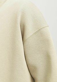 Material de sudadera beige con una textura suave. Presenta un cuello redondo, un corte relajado y hombros ligeramente caídos. Diseño sencillo y sin adornos.