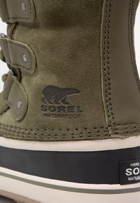 Nahaufnahme eines olivgrünen Wildlederschuhs von Sorel, wasserdicht, mit einer schwarzen und weißen Gummisohle und gestickten Lederdetails.