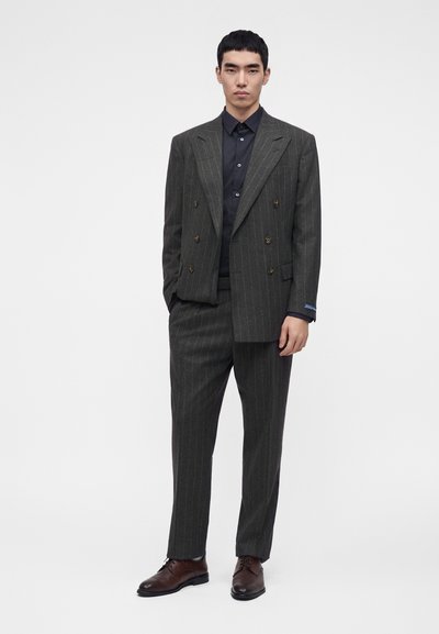 Homme debout portant un costume croisé gris foncé à fines rayures avec une chemise noire et des chaussures marron sur un fond blanc uni.