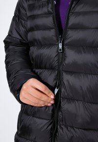 Chaqueta acolchada negra con diseño de rombos, cierre de cremallera y textura brillante prominente. Una mano sostiene el tirador de la cremallera, mostrando los detalles del herraje metálico.