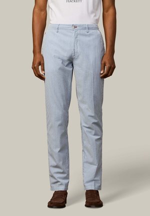 Pantalon slim rayé bleu clair et blanc porté avec une chemise blanche et des mocassins en daim marron sur un fond neutre.