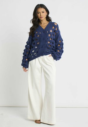 Woman with long dark hair wears oversized navy blue open-knit cardigan and wide-leg white pants, standing against plain background.